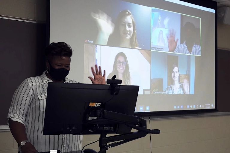 A teacher wearing a mask in a classroom waves to students seen on projector screen.