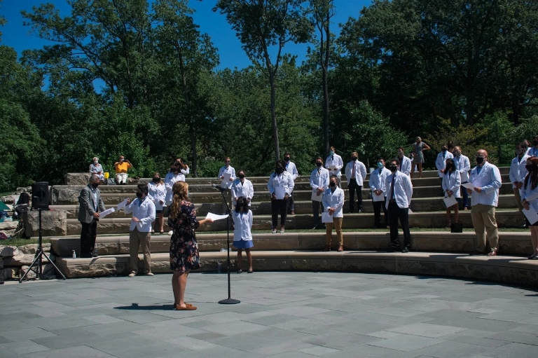 People wearing white medical coats face a speaker during a ceremony