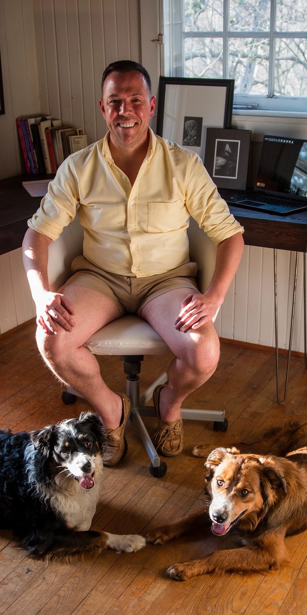 A man sits near his home computer station with two dogs at his feet
