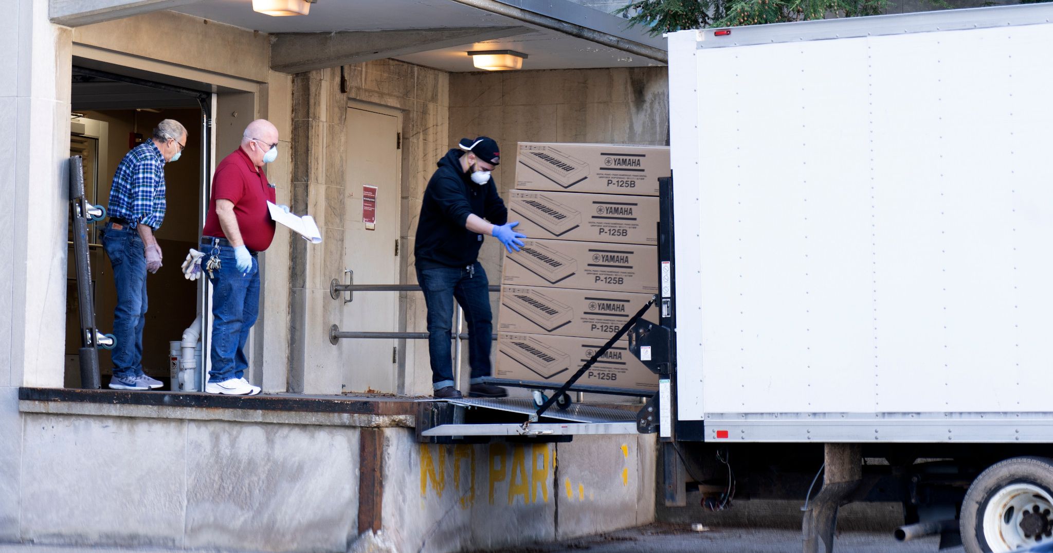 Gloved and masked workers move boxed Yamaha digital pianos from a truck to a loading dock.