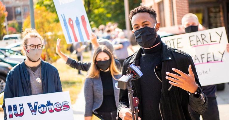 A student in a face mask speaks into the microphone while other students hold up signs.