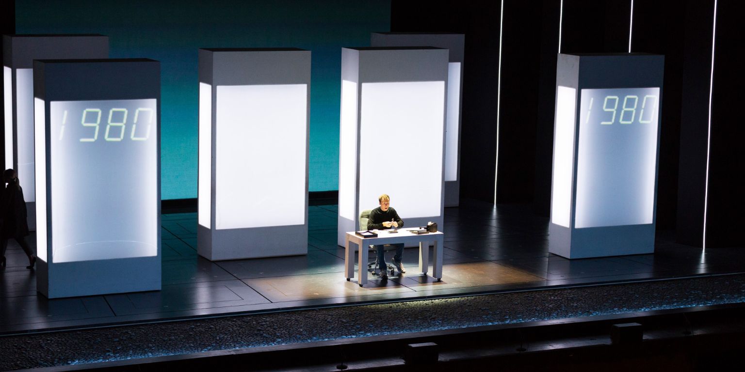 A man sits at a desk in front of screens that read 1980