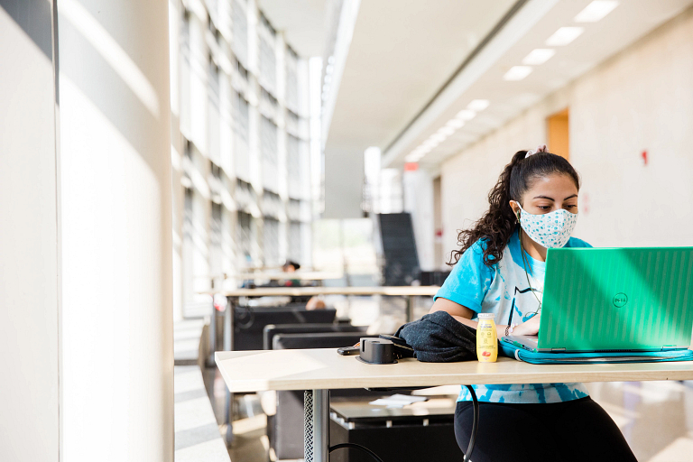 A student wearing a mask sits at a table working on a laptop