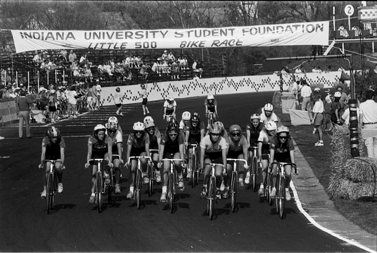 Cyclists on bikes line up at the starting point of a race with a banner hanging above them.