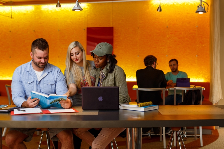 Three students look at a book together at a table.