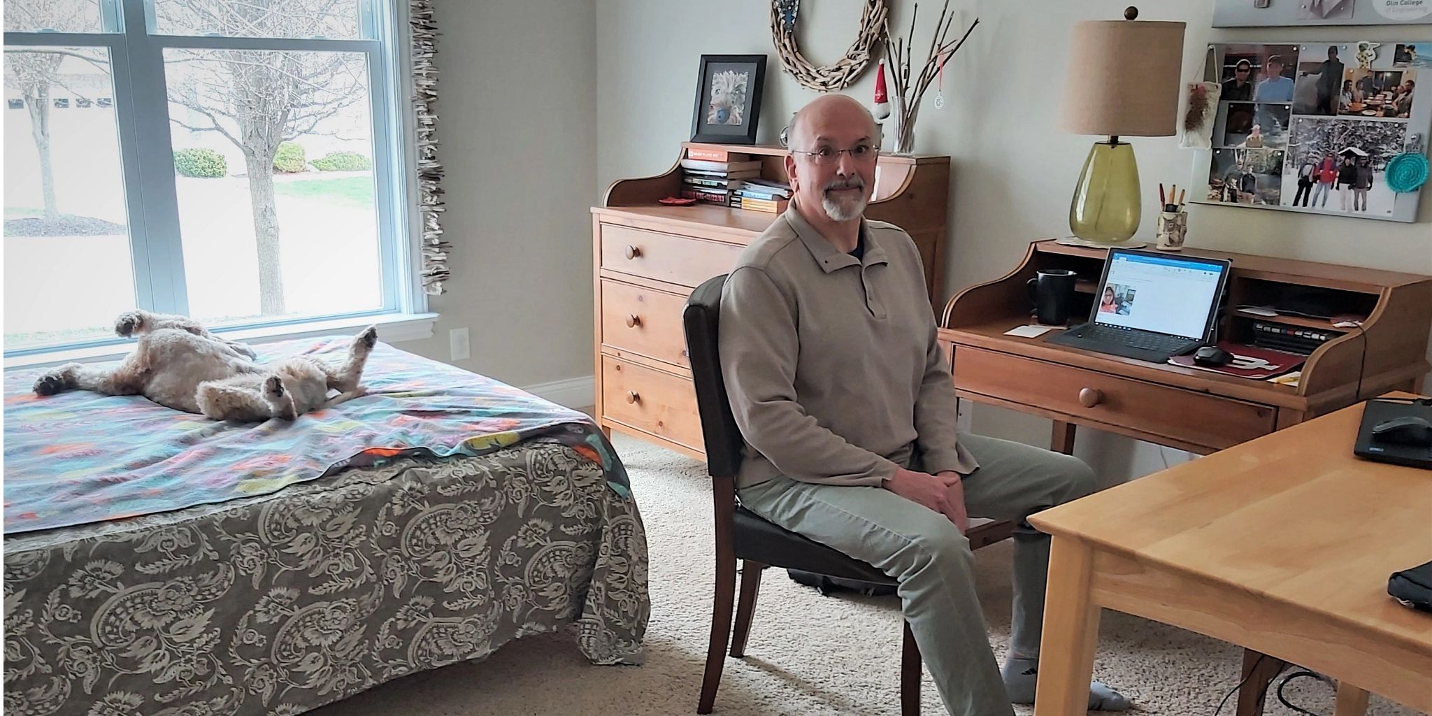 A man sits at a home office space in his bedroom while a dog lies on the bed behind him