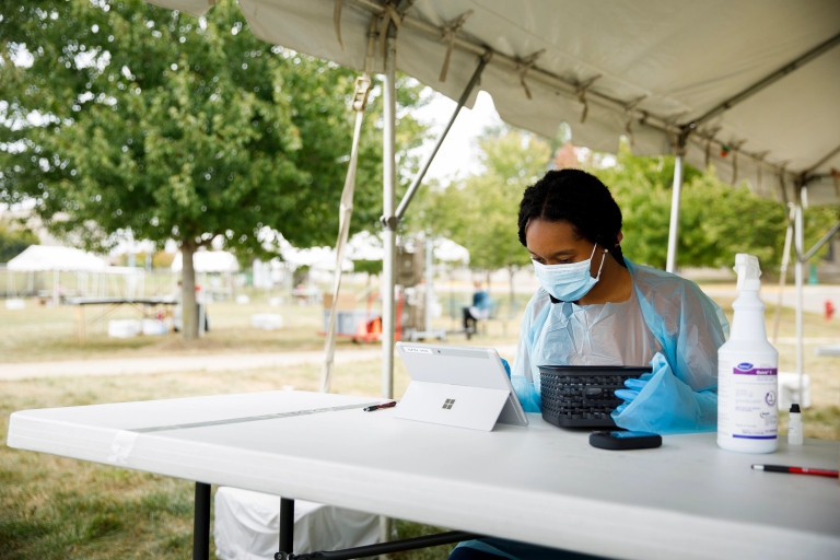 A student in gloves and mask using a tablet computer under a tent outdoors.