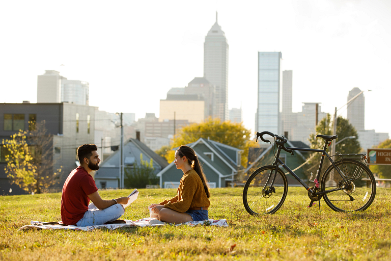Two students sit in the grass next to a bicycle with Indianapolis skyline in background.