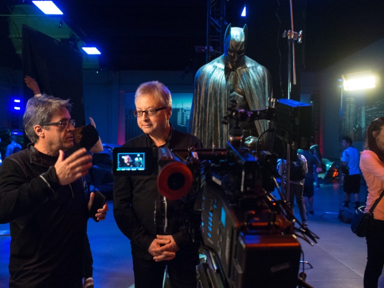 Angelo Pizzo and Michael Uslan stand in front of a camera with a Batman looming in the background