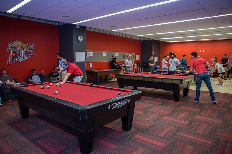 The game room shows people playing pool at two pool tables with other activities in the background.