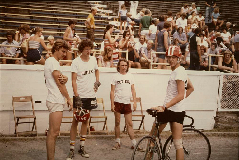 Four men in shorts and T-shirts stand together outdoors on a bicycle track.