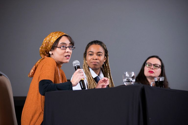 A woman speaks into a microphone while sitting at a table with two other women.