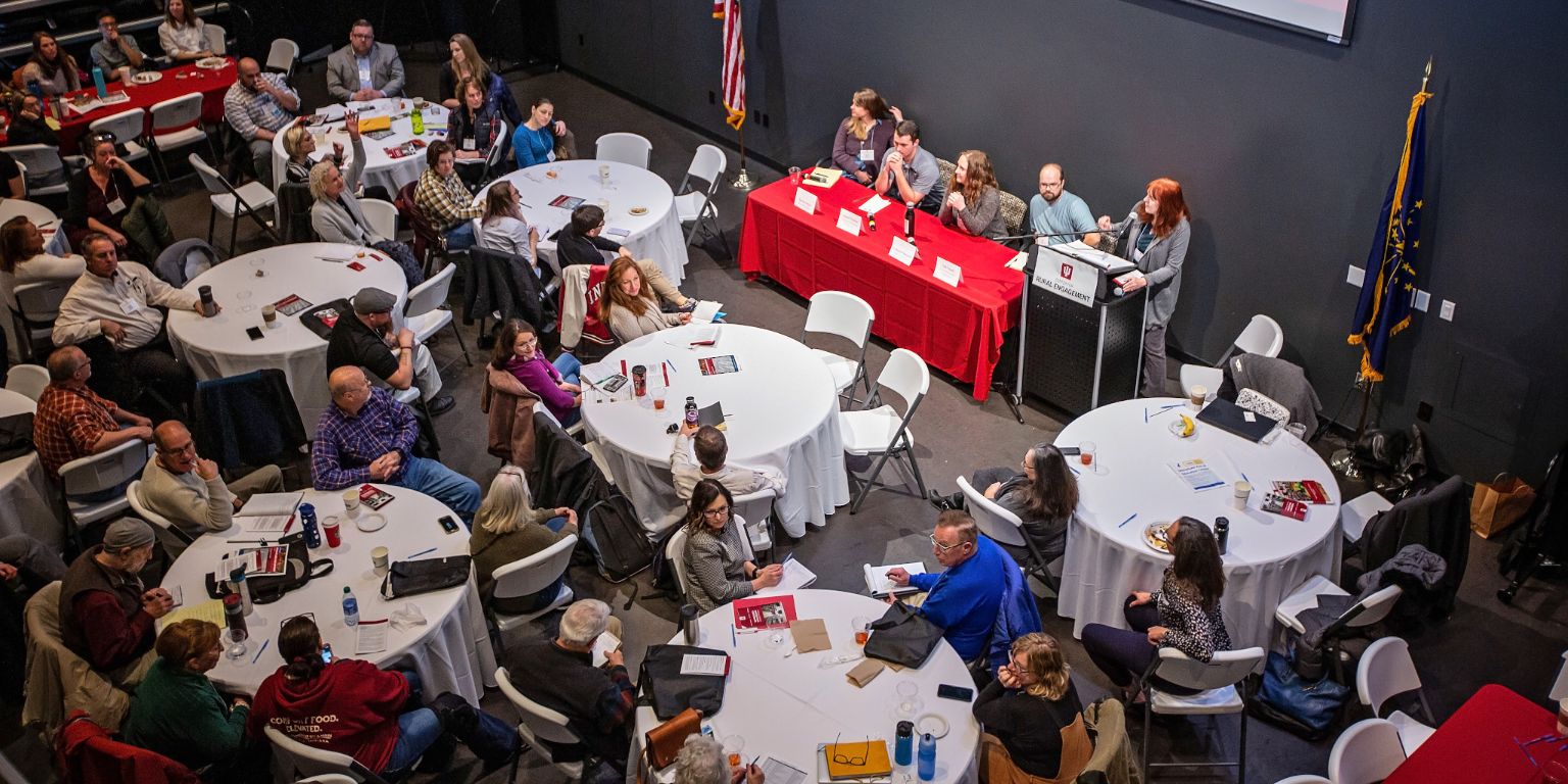 Conference attendees sit at round tables watching a panel discussion