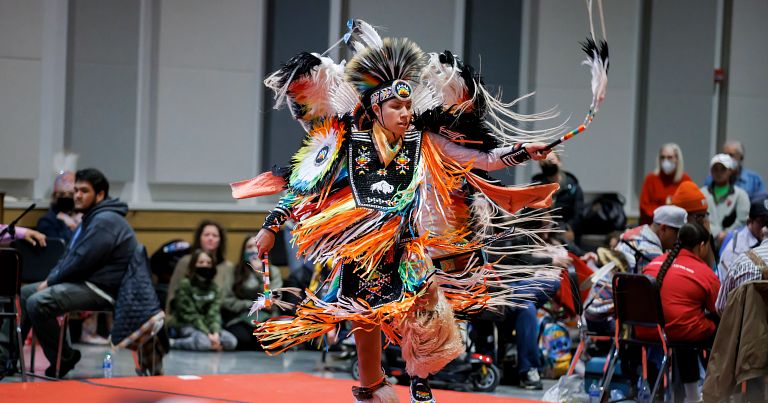 An indigenous artist in colorful clothing performs a dance.
