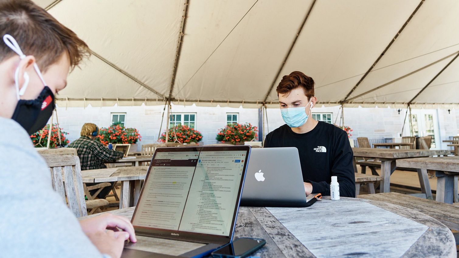 masked students work on laptops while seated under a tent