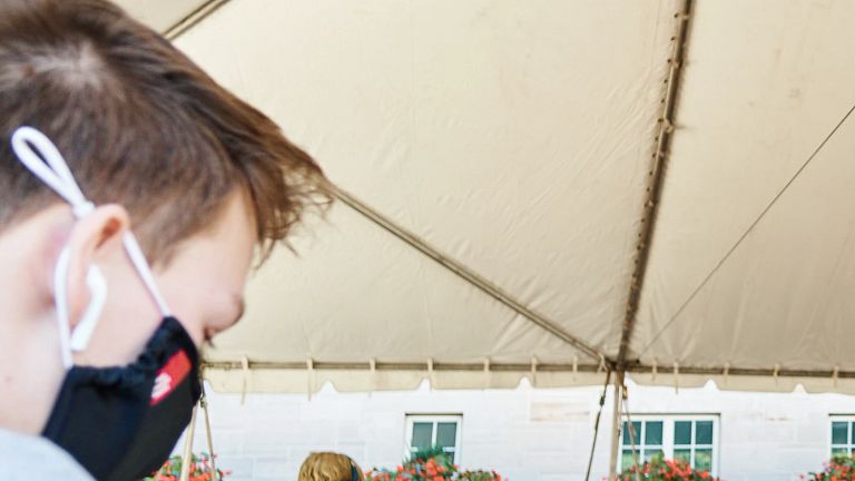 masked students work on laptops while seated under a tent