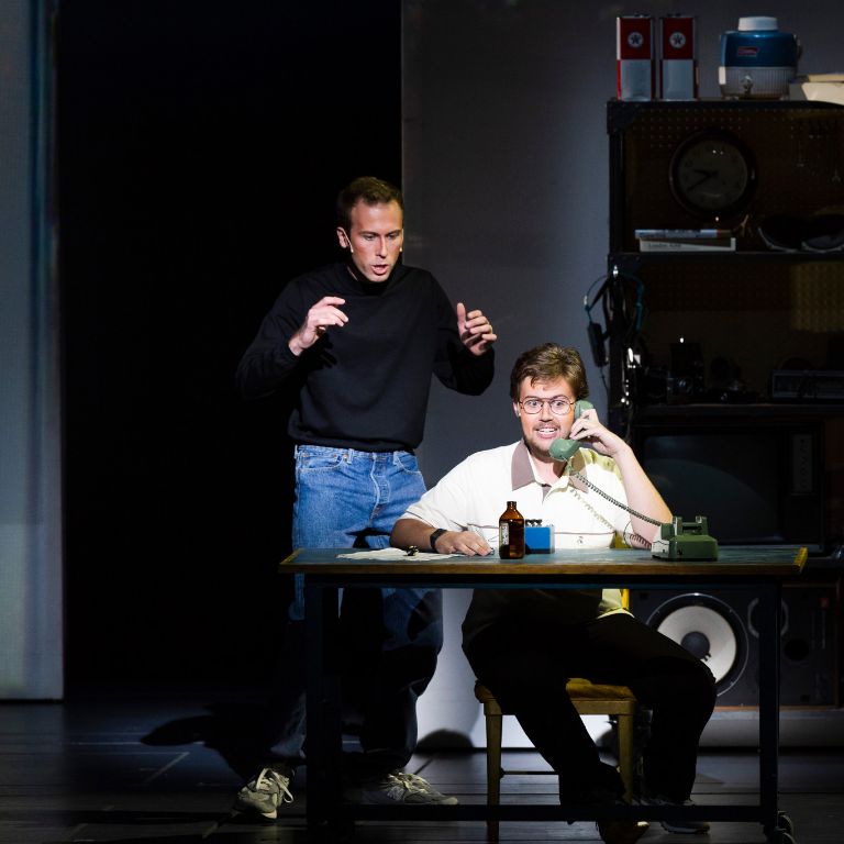 A man sits at a desk on a phone and another man stands behind him