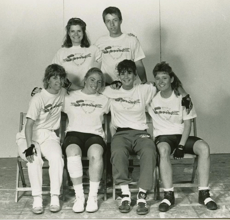 Four women seated in chairs smiling. A woman and man stand behind them.