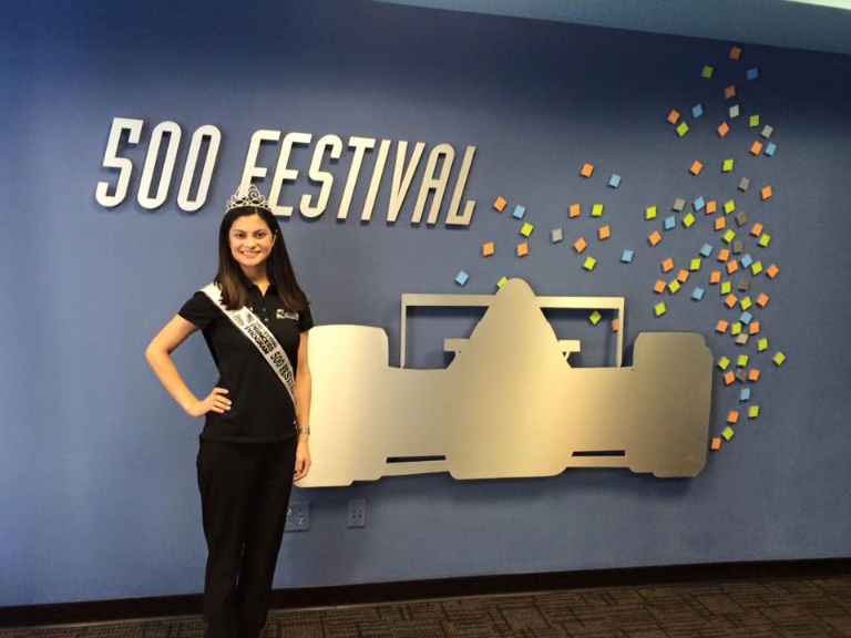 Amna Sohail stands in front of a blue wall with art depicting an Indy car for the 500 Festival.