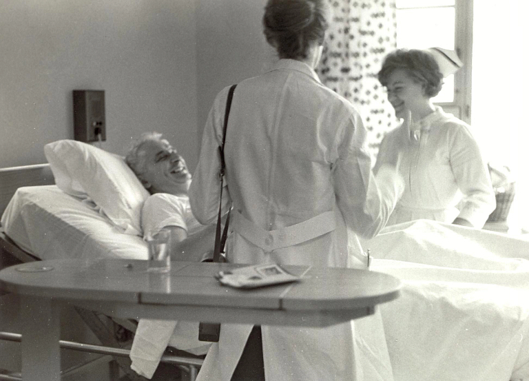 Two nurses stand next to a man in a hospital bed.