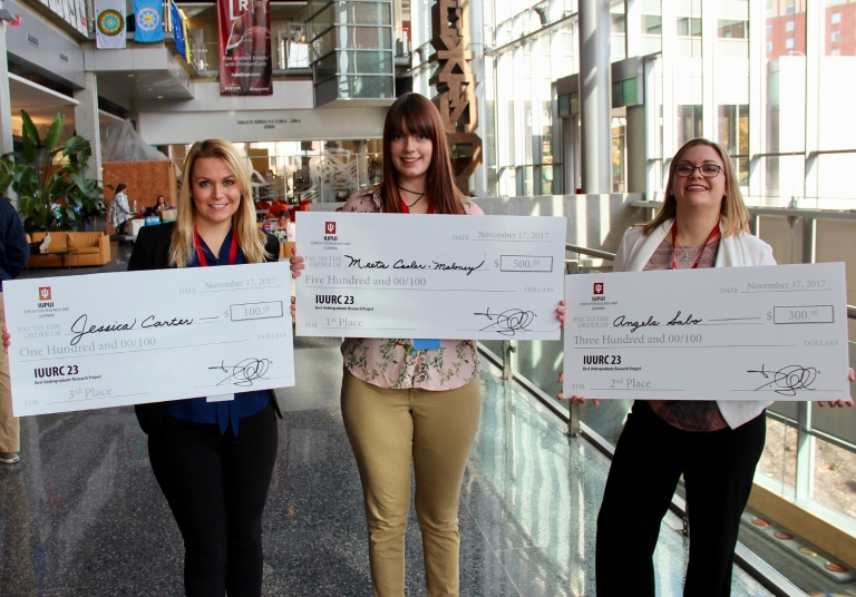 Three women hold giant checks from winning posters in the IU Undergraduate Research Conference.