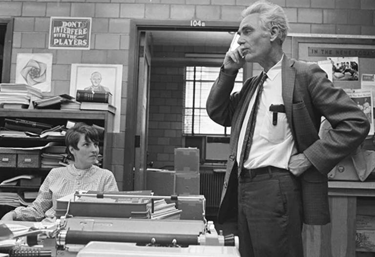 A black and white photo of a woman looking at a man standing up talking on the phone