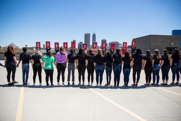 Students on top of sports garage hold signs with Indianapolis skyline in the background.