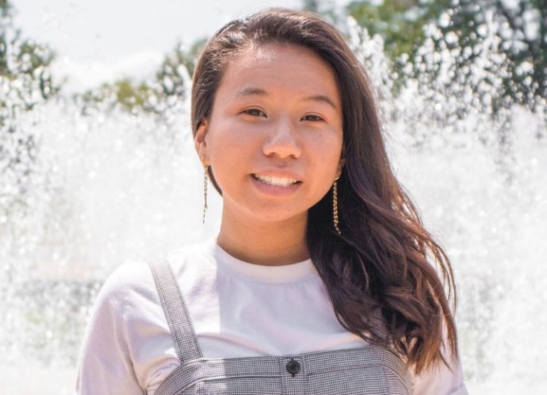 Thi Pham smiles and poses for a photo in front of a large water fountain on a sunny day.
