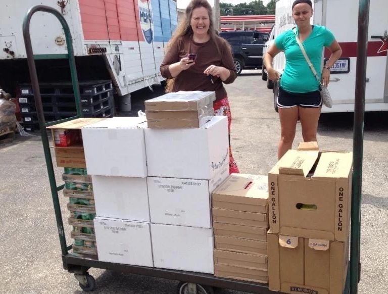 Two women stand by a large dolly stacked with canned food