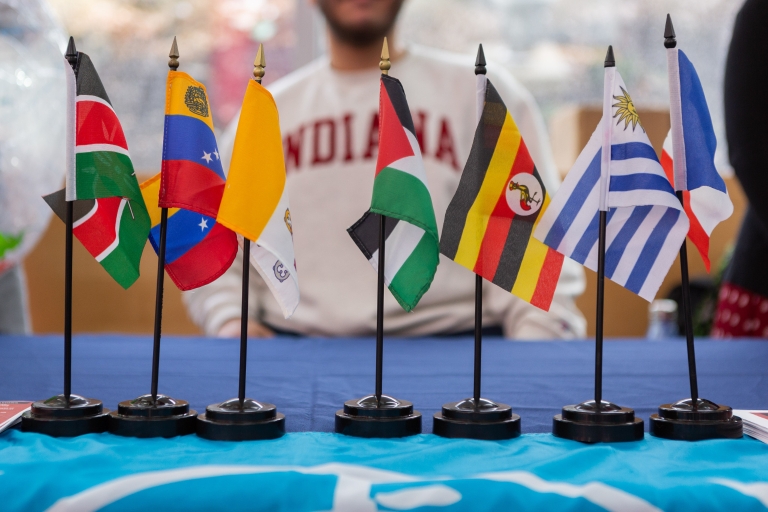 Flags from countries around the world are displayed on a table with an IU student in the background