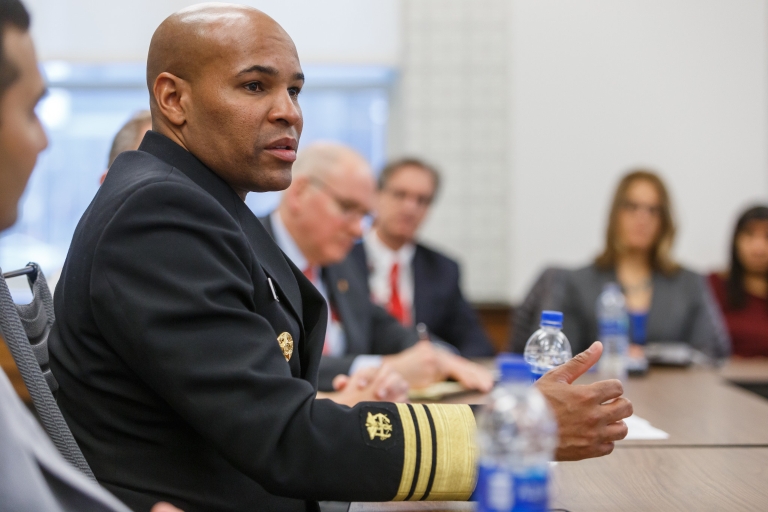 U.S. Surgeon General Jerome Adams speaks to a group of people while seated at a table.