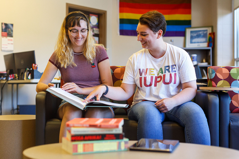 A student and mentor sit on the couch in the LGBTQ+ Center, looking at a book.