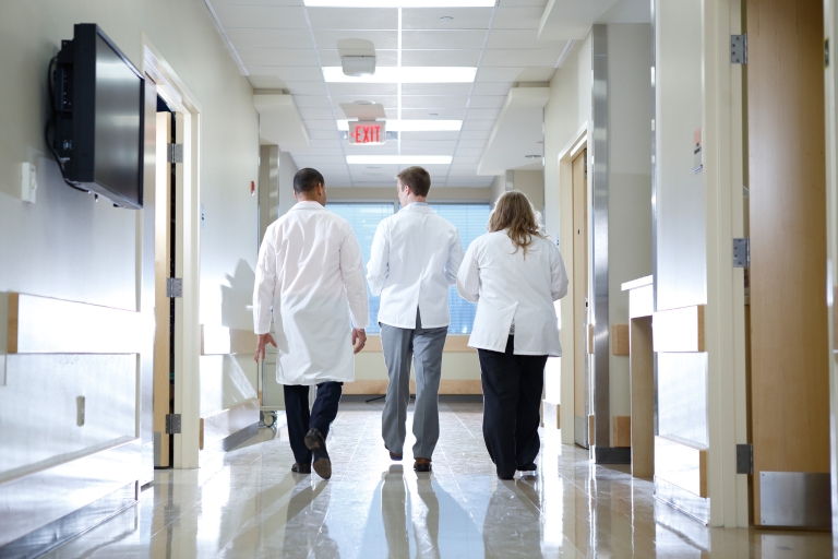 Three people in white lab coats walk down a hall of a hospital. 