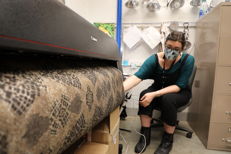 A woman sits next to a digital printer as it prints a strip of fabric with a python pattern