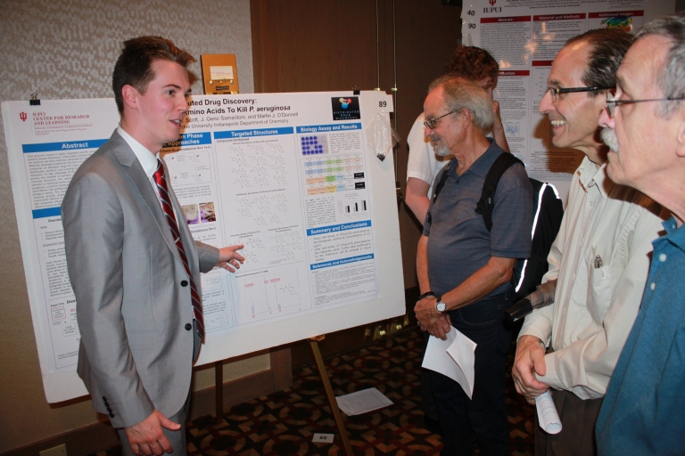 Isaac Lamb points to a large poster while explaining his research to three professors.