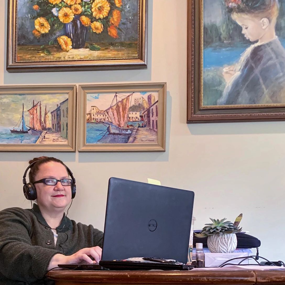 A woman works on her laptop sitting next to pieces of artwork mounted on a wall.