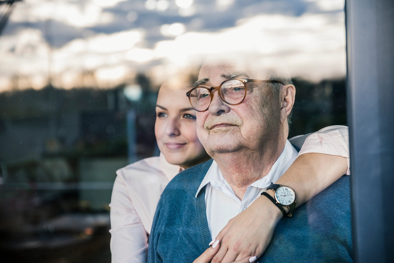 Portrait of a young woman with her arms around a senior man at the window