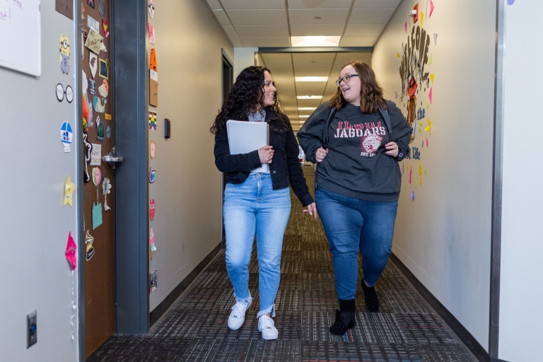 Two women walk down a hallway while chatting.