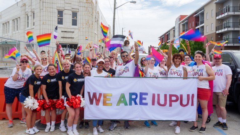 A large group of people hold pride flags and a banner that reads We are IUPUI