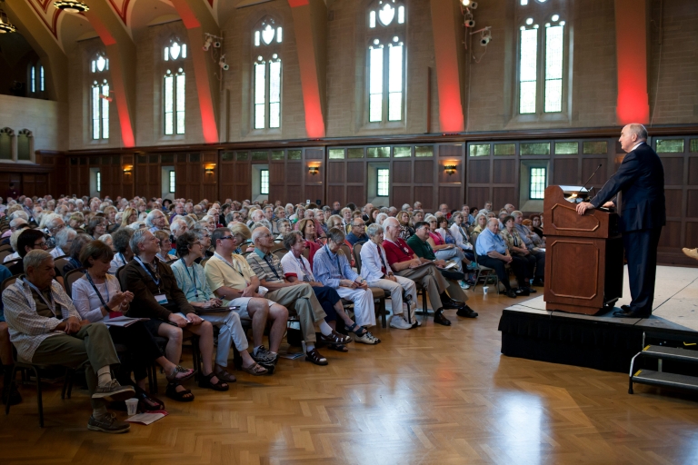 President Michael A. McRobbie stands on a podium to speak to a crowd of attendees in Alumni Hall.