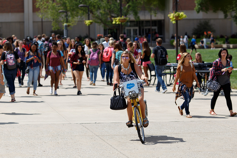 Woman rides a bike through campus while students walk behind her.